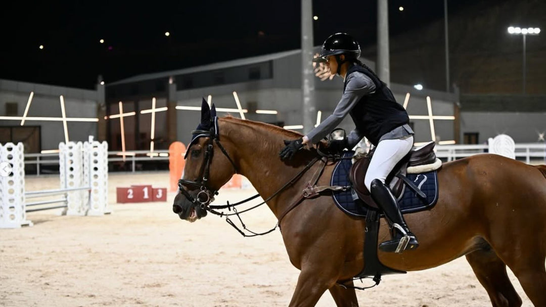 Female rider in helmet, boots and white breeches on a bay horse in a floodlit show jumping arena
